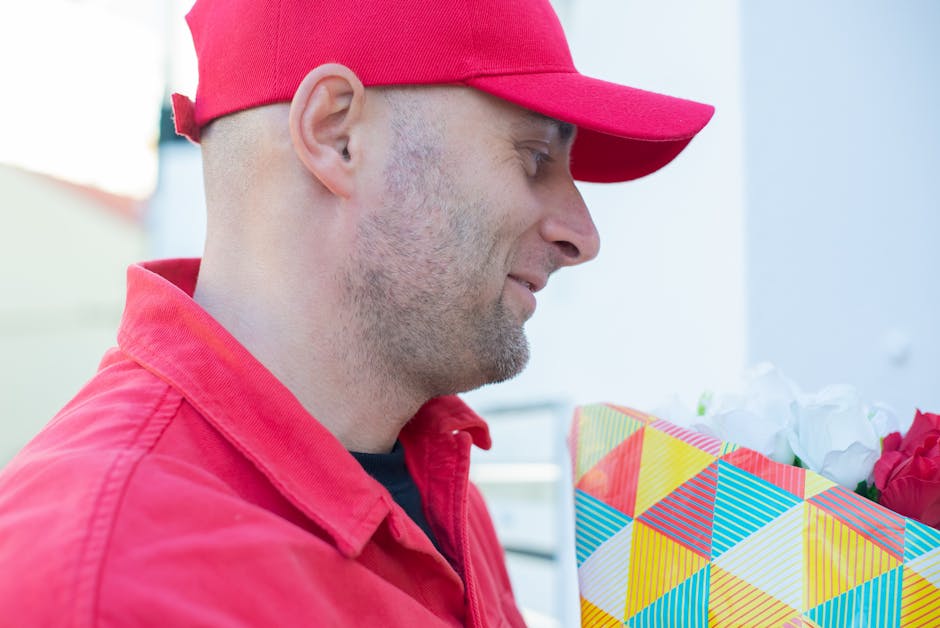 A man dressed in a red cap and red jacket is holding a colorful, geometric-patterned gift box filled with pink and white flowers. The arrangement features a mix of roses and other blooms, with visible