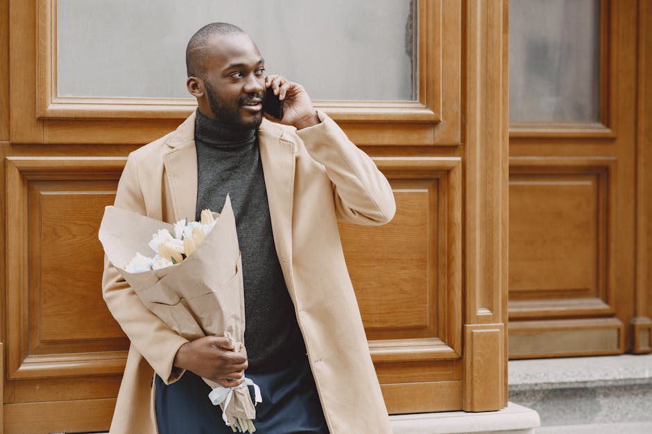 A young man with dark skin, short hair, and a neatly trimmed beard is standing outside in front of a wooden-paneled building, holding a bouquet of fresh white roses wrapped in brown paper. He is weari