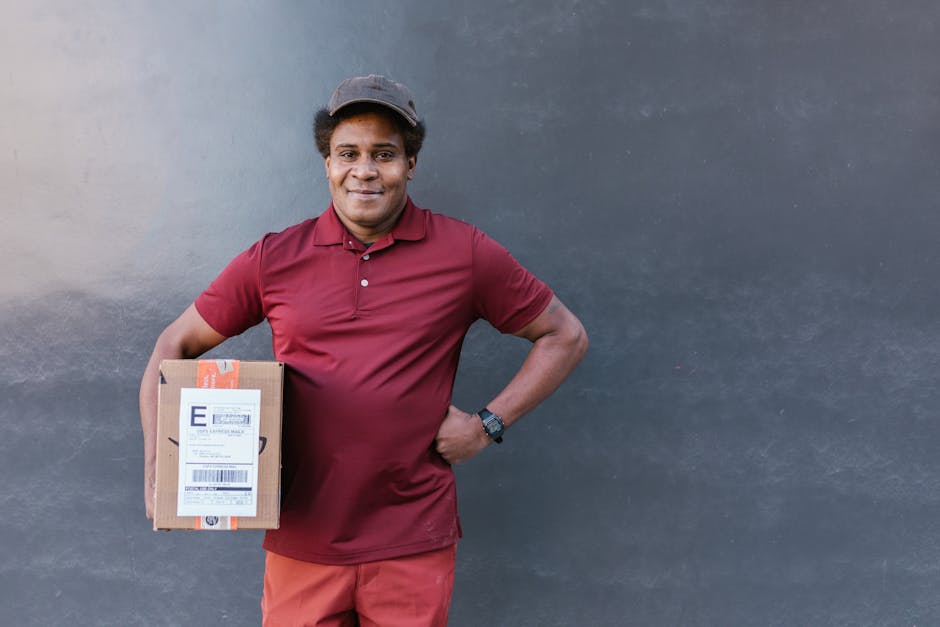 A male delivery person standing against a plain gray wall, smiling and holding a cardboard parcel with a printed label, dressed in a maroon polo shirt, maroon trousers, and a gray cap. He appears read