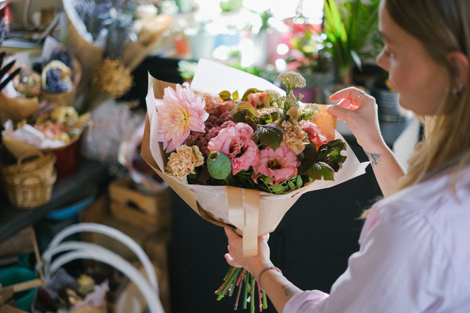 A woman holding a bouquet of fresh flowers inside a floral shop, arranged with large pink dahlias, pale pink lisianthus, beige carnations, and lush green foliage. The bouquet is wrapped in light brown