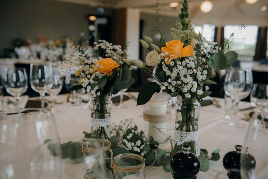 A floral arrangement featuring small bouquets of yellow roses, white roses, and white baby's breath, complemented by green foliage. The flowers are displayed in three clear glass jars decorated with d