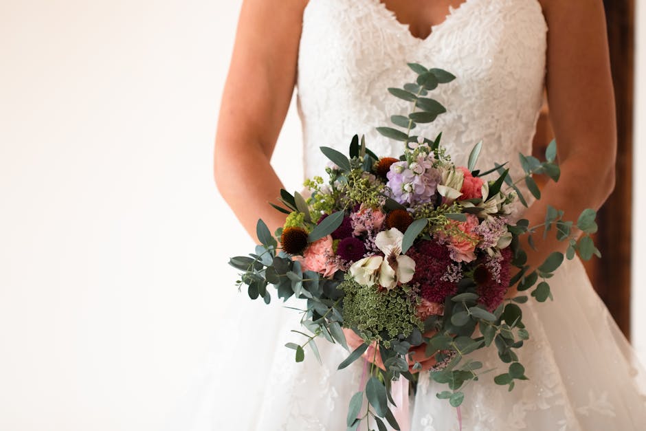 A bride dressed in a white lace wedding gown is holding a large, rounded floral bouquet in both hands. The bouquet features a variety of fresh flowers, including soft pink, deep red, and white blooms,